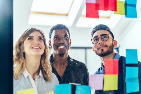 Multicultural group of creatie male and female colleagues reading notes written on colorful stickers standing behind glass wall in office.Positive employees collaborating on task using sticky papers