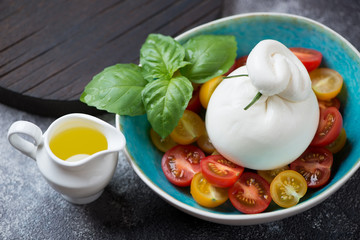 Burrata cheese with cherry tomatoes and fresh green basil in a turquoise bowl, studio shot over light-grey stone background
