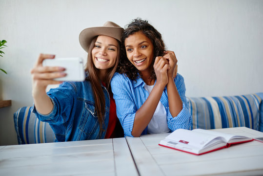 Cheerful multiracial hipster girls in trendy clothes posing for selfie on smartphone camera in cafe,smiling woman taking picture with her african american friend for share in networks via cellphone.