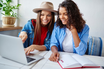 Cheerful multiracial female student making homework together making research on netbook having fun during working process,smiling women browsing web store page on laptop computer talking about items .