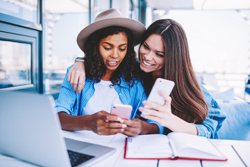 Funny female friends in trendy outfit synchronizing files with mobile phones sending common photos...