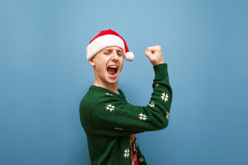 Portrait of joyful young man in christmas clothes, wearing santa hat and sweater, rejoices in victory on blue background. The guy won at Christmas, he is happy. Isolated, New Year.