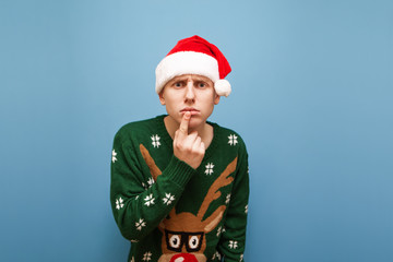 Pensive young man stands against a blue background, looks into the camera and thinks, wearing a Christmas sweater and hat of santa claus, isolated. Portrait of a thoughtful guy in Christmas.