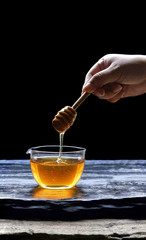 Focus on woman hand holding wooden honey dipper stick and dipping from glass cup on black slate...