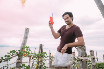 A young man stands by the sea on a wooden bridge with a cocktail.