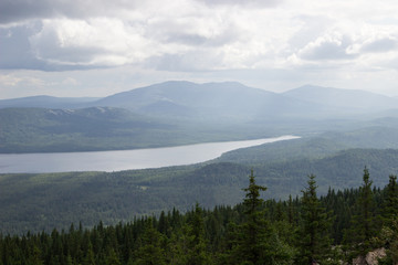 Beautiful mountain landscape with a lake in the middle of a spruce forest on a cloudy day, a photo from a height in the foreground bright spruce trees with fading colors as they move away