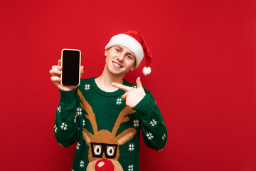 Happy guy in santa hat and christmas sweater holds smartphone in his hand and points finger at the screen and looks in camera,red background. Smiling man showing thumbs up on black smartphone screen