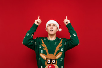 Shocked handsome young man in christmas clothes and santa hat on red background looking and pointing upwards to empty space. Surprised guy shows up on copy space. Isolated.