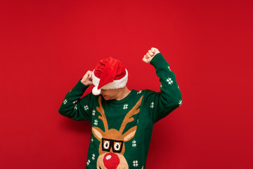 Cheerful young man in santa hat and green sweater dancing on red background. Cheerful guy having...
