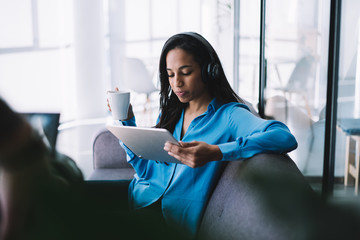 Ethnic woman browsing on tablet and listening to music while drinking coffee