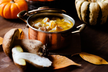Delicious hot porcini soup in copper pan. Tasty seasonal autumn meal. Fresh mashrooms illustrate ingredients. Concept of homemade food. Closeup, dark wooden background. Pumpkin on the back