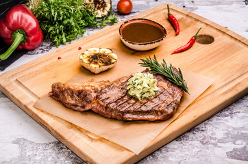 Medium rare grilled beef steak on a rustic cutting board with rosemary and spices, bbq sauce, red chili pepper, fried garlic, vegetables at a table restaurant background.