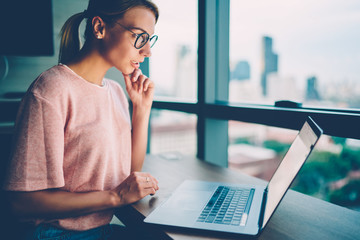 Pensive smart young woman in eyeglasses reading last news in social networks at laptop gadget...