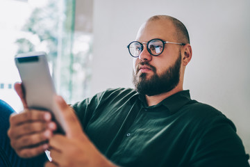 Pensive caucasian male in spectacles reading e book spending time on hobby at home interior, serious bearded hipster guy browse web page on digital tablet making booking sitting at apartment