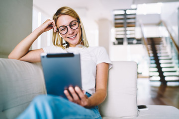 Positive blonde young woman in optical spectacles watching funny tutorial on touch pad resting on couch at home.Cheerful blogger in earphones laughing during reading comments from followers on tablet - obrazy, fototapety, plakaty