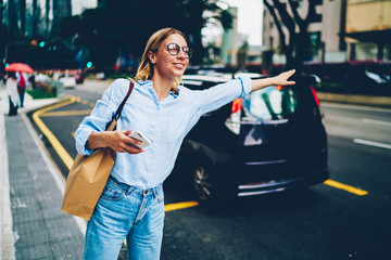 Pretty hipster girl in eyewear raising hand standing on road waving for take a cab in downtown, young woman hailing for taxi waiting for transport in city center holding smartphone and shopping bag