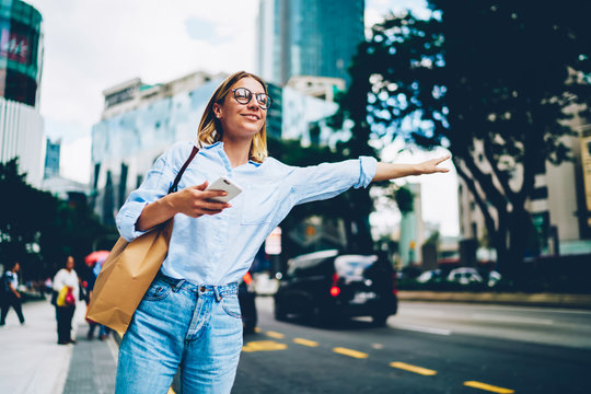 Smiling female traveler in trendy casual wear raising hand calling taxin standing on road in city on urban background, young woman waiting for cab for transporting in megalopolis waving on avenue