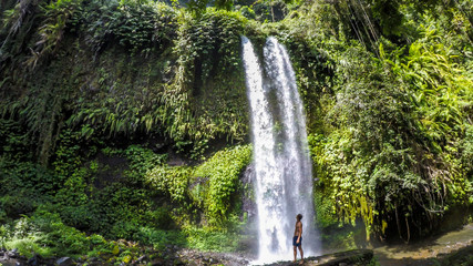 Fototapeta na wymiar A man in swimsuit standing under two levelled waterfall in Lombok, Indonesia. Tiu Kelep Waterfall is surrounded by lush green plants from each side. Long and powerful waterfall. Beauty of the nature.