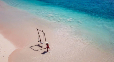 Fototapeta na wymiar A girl swinging on a swing placed on the seashore of Pink Beach, Lombok, Indonesia. The swing has simple wood construction. Waves wash the pillars of it. In the back there are few boats. Drone capture