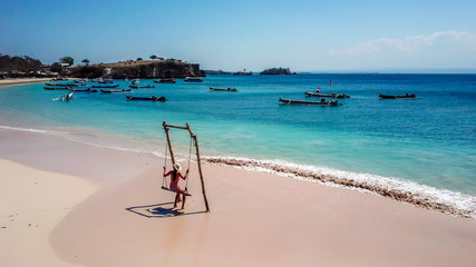 A girl swinging on a swing placed on the seashore of Pink Beach, Lombok, Indonesia. The swing has simple wood construction. Waves wash the pillars of it. In the back there are few boats. Drone capture