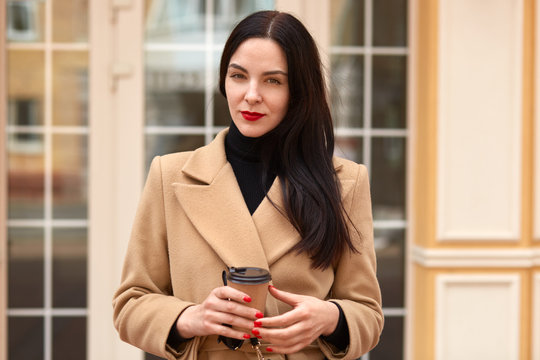 Horizontal image of serious confident businesswoman holding papercup of coffee, looking directly at camera, posing over glass door outside, wearing beige coat and black sweater, being at lunch.