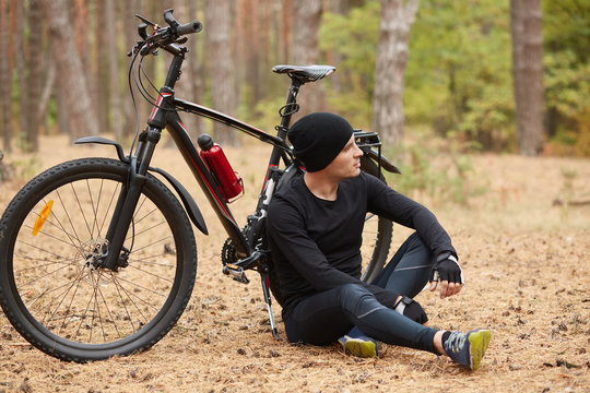 Outdoor picture of athletic strong sportsman wearing black shirt, sport pants, hat and trainers, leaning on bicycle, looking far ahead, having training around nature. Nature and people concept.