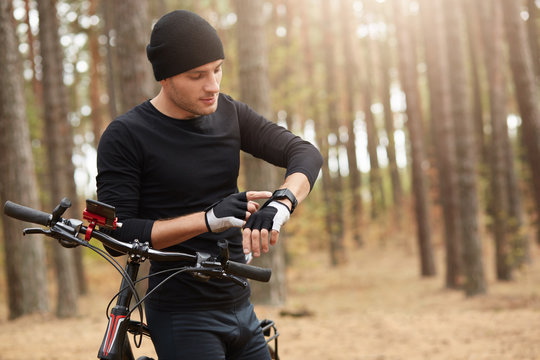 Horizontal image of athletic sportsman using his smartwatch, looking at its screen, touching it, having bicycle walk, leaning on bike, wearing black sportwear, being in autumn forest. Sport concept.
