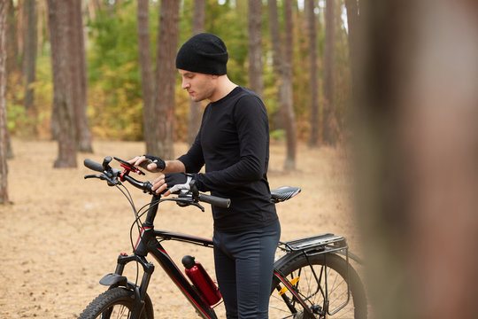 Picture of attentive adventurous man standing in middle of forest, holding his bicycle, looking at smartphone screen, looking for way back, using GPS, choosing shortest way. New technologies concept.