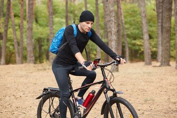 Fototapeta premium Confident handsome cyclist riding his bike in autumn forest, following his way, having map on his smartphone, wearing black sportswear, having blue backpack, spending time around nature alone.