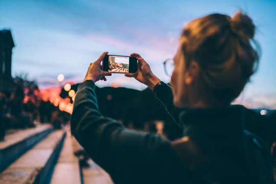 Back view of hipster girl taking pictures of evening city via smartphone application connected to 4g for using cellular online app, female generation photographing landscape during sightseeing