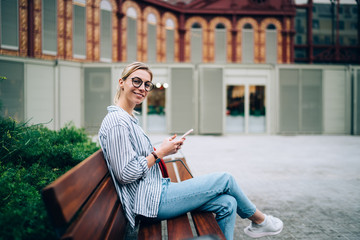 Smiling stylish woman using phone on bench and looking at camera