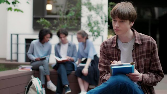 Medium shot of casual student boy writing notes in notebook with group of students on background in university campus outdoor