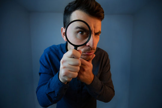 Surprised Young man student holding magnifying glass looking to camera with a pensive emotion isolated over grey  background. Science and curiosity concept.