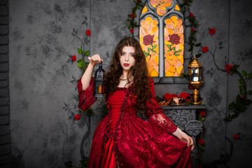 A beautiful girl in a magnificent red dress of the Rococo era stands against a fireplace, a window and flowers with a lamp with candles in her hands.