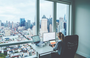 Rear view female employee working on PC computers with financial information on screen sitting at office table with downtown skyscrapers view behind window. Successful businesswoman at her workspace