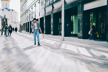 Stylish young man strolling outdoors with paper bags in hands near shopping center while reading incoming message on smartphone device.Hipster guy customer with packages dialing number on mobile phone