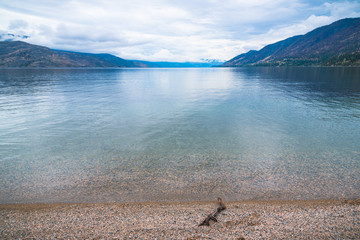 View of pebble beach, calm lake, mountains, and overcast sky at Antler's Beach, Peachland, British...