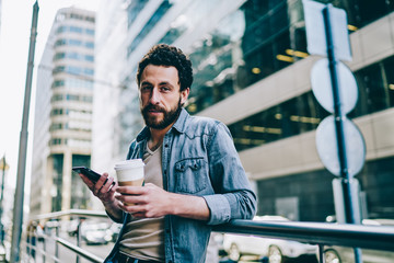 Portrait of bearded young man dressed in denim wear looking at camera while holding coffee to go and smartphone in hands standing on balcony.Hipster guy with cup of caffeine updating app on cellular