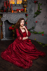 A beautiful girl in a magnificent red dress of the Rococo era sits on the floor near a fireplace and flowers.