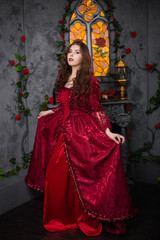 Beautiful girl in a magnificent, red rococo dress. Against the background of the fireplace, window, and flowers.