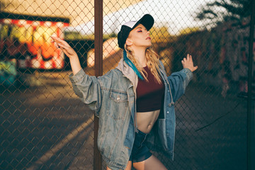 Young attractive woman in jeans jacket, shorts, red top and trucker hat posing over metal fence and graffiti wall over background in a city
