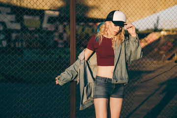 Young attractive woman in jeans jacket, shorts, red top and trucker hat posing over metal fence and...