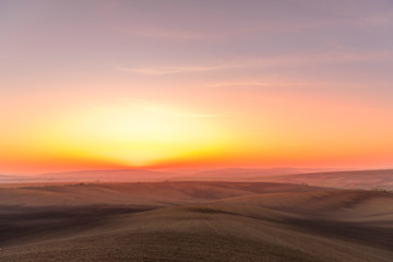 Fototapeta na wymiar The sun caught during the last moment before setting beyond the horizon with view of a field covered with fog and the farms and fields undulating in the region of South Moravia is also called Tuscany