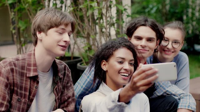 Close up shot of young cheerful multinational students happily taking selfie on cellphone resting in university campus outdoor