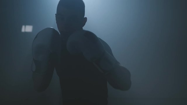 Male boxer punching to camera. Silhouette of young boxer practicing boxing punches looking at camera in dark smoky gym. Close-up in 4K, UHD