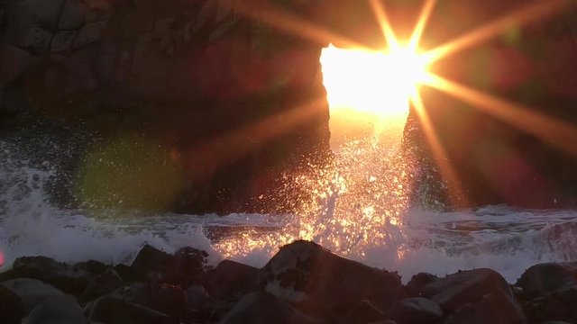 Slow motion waves crash over smooth stone at Pfeiffer Beach as a sunstar forms against the hard rock sea stack in this amazing natural wonder from Western California on the Pacific Ocean coast. 