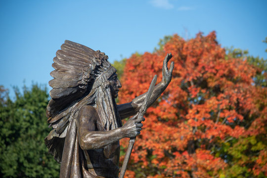 The Statue Of Native American Indian In A Head Dress Stands Out Among The Changing Leaves Of The Trees During A Sunny Day In Autumn