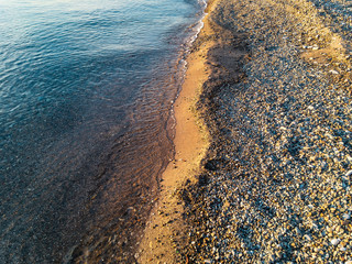 The coastline is covered with pebbles and sand with a sea wave of clear water running onto it, illuminated by yellow sunlight at sunset on an autumn evening.
