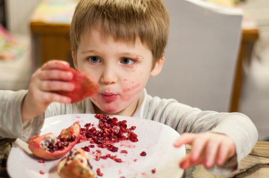 Portrait Of Blonde Baby Boy Eating Pomegranate. Breakfast At Home In The Kitchen. The Boy Smeared In Red Juice Of Fresh Fruit. Healthy Nutrition, Natural Fruits And Vitamins. Funny Dirty Kid.