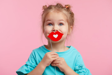 Funny smiley girl face on the background of a bright pink wall. Child girl with paper accessories, paper lips on a stick.